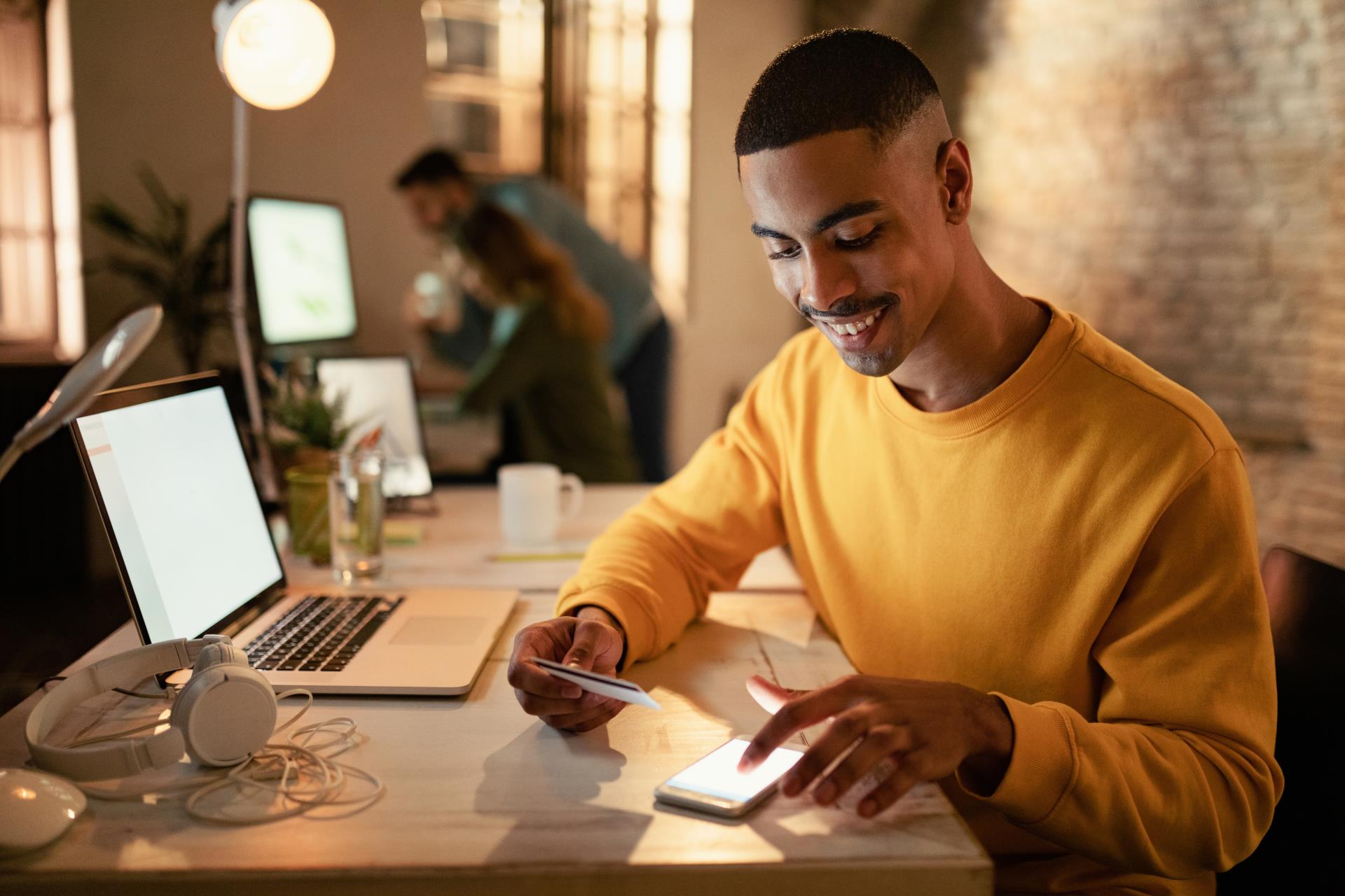 happy-african-american-entrepreneur-using-mobile-phone-credit-card-from-online-payments-while-working-night-office