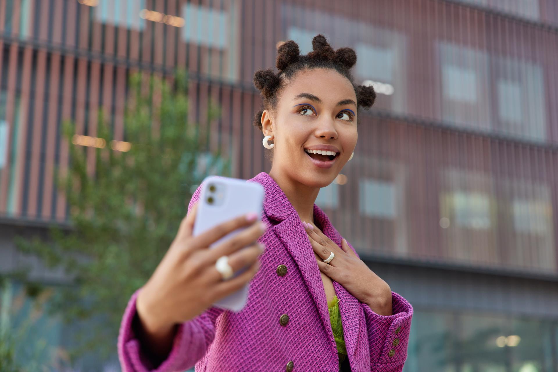 happy-dreamy-woman-with-funny-hairstyle-makes-photo-herself-wears-fashionable-pink-jacket-looks-somewhere-positively-poses-outdoors-against-blurred-background-technology-urban-lifestyle