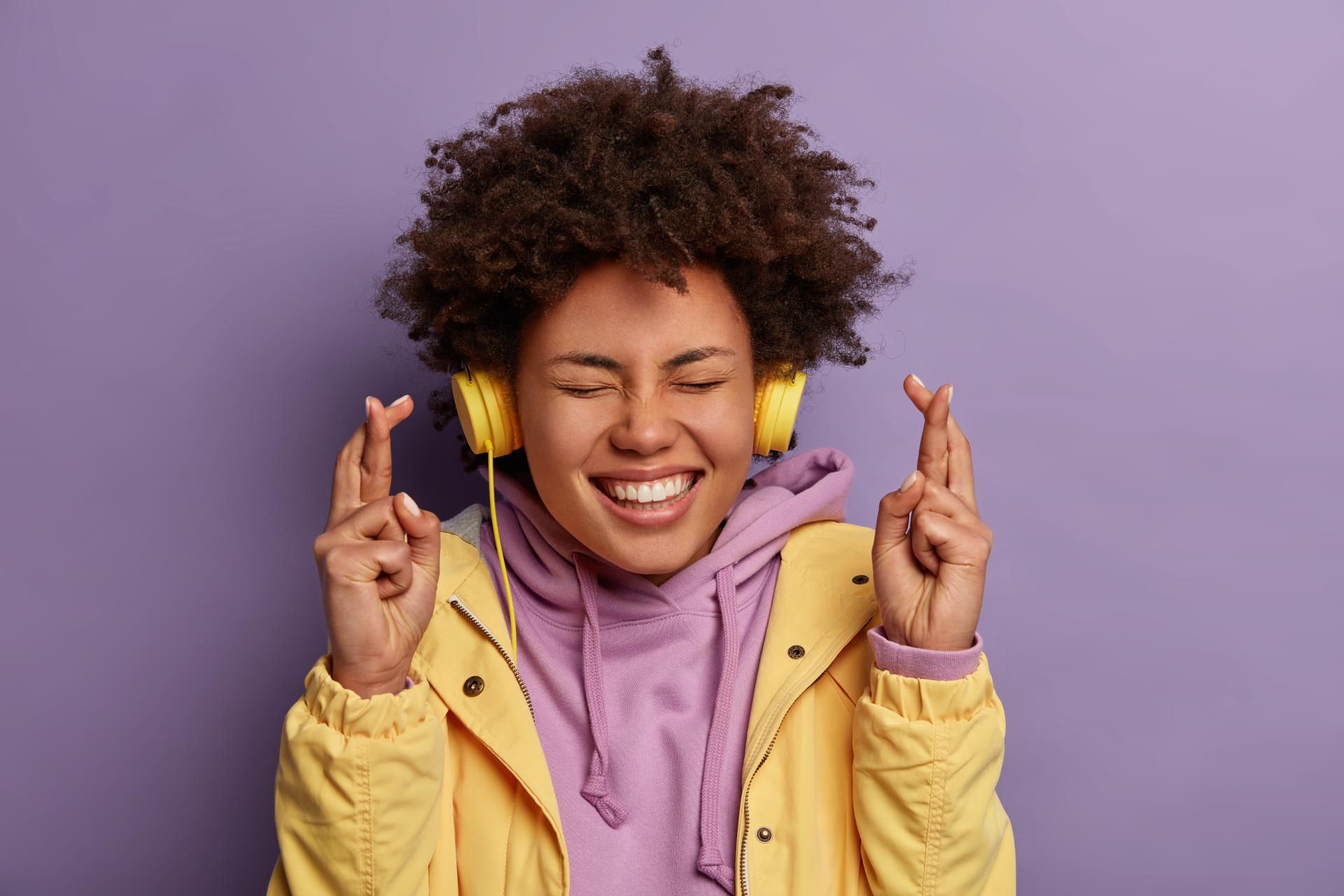 wishful-happy-woman-with-natural-curly-hair-anticipates-relish-good-news-crosses-fingers-smiles-broadly-awaits-dream-come-true-wears-stereo-headphones-listens-pleasant-music