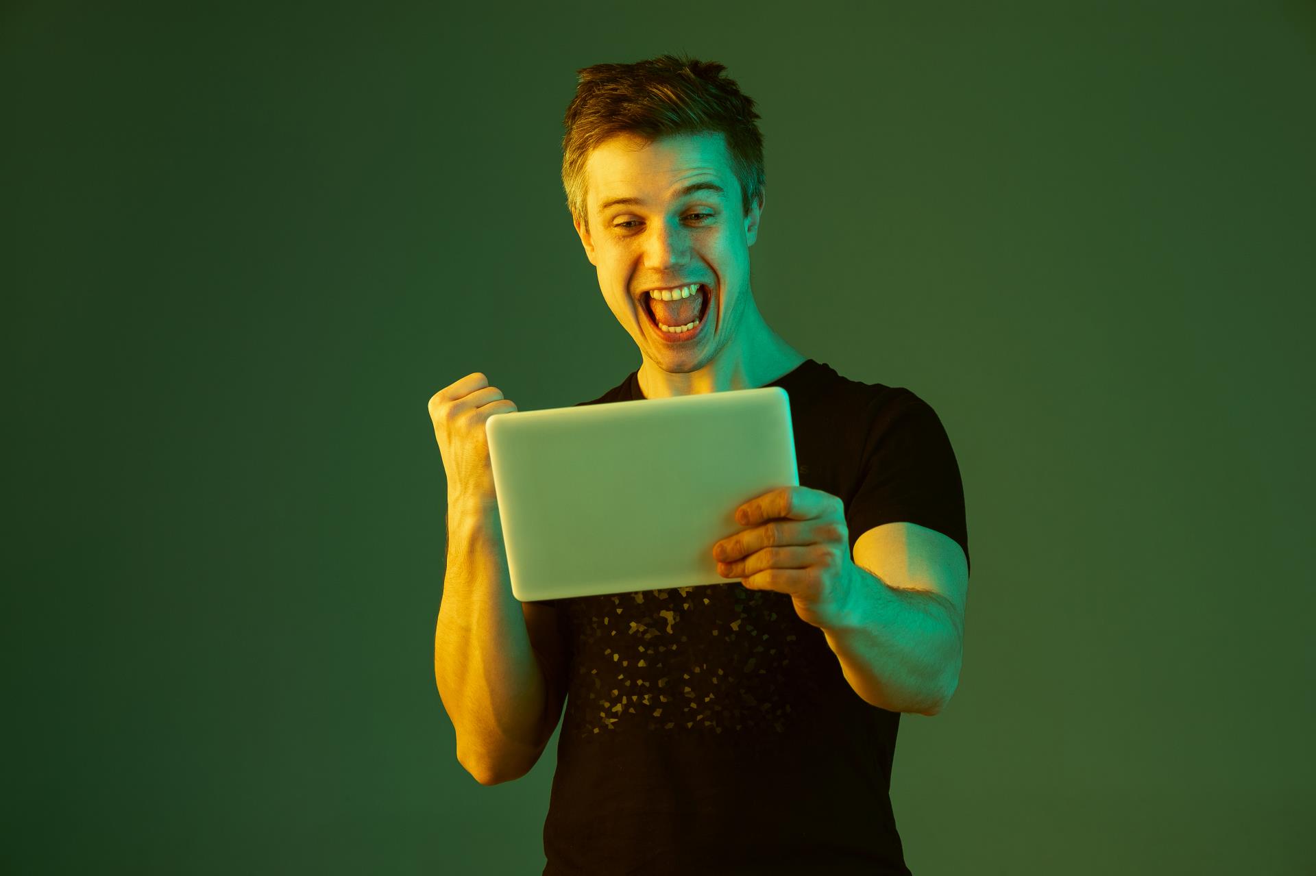 holding-tablet-celebrating-win-bet-game-caucasian-man-s-portrait-green-studio-background-neon-light-beautiful-male-model-concept-human-emotions-facial-expression-sales-ad